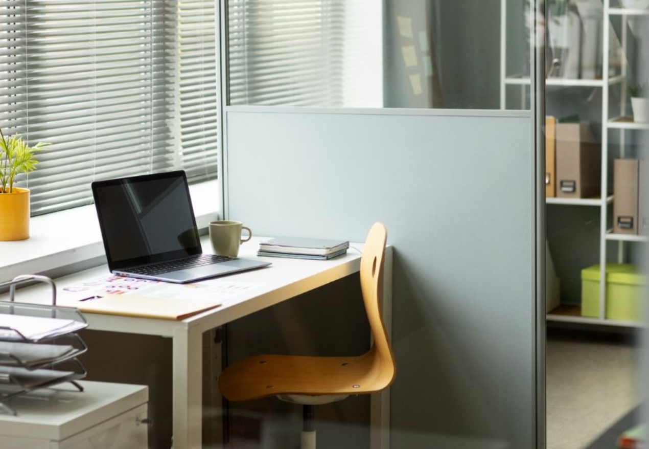 A modern office cubicle with a wooden chair, laptop, coffee mug, and a small potted plant on the desk. Sunlight filters through the blinds, creating a bright and welcoming workspace. Shelving with files and boxes is visible in the background.