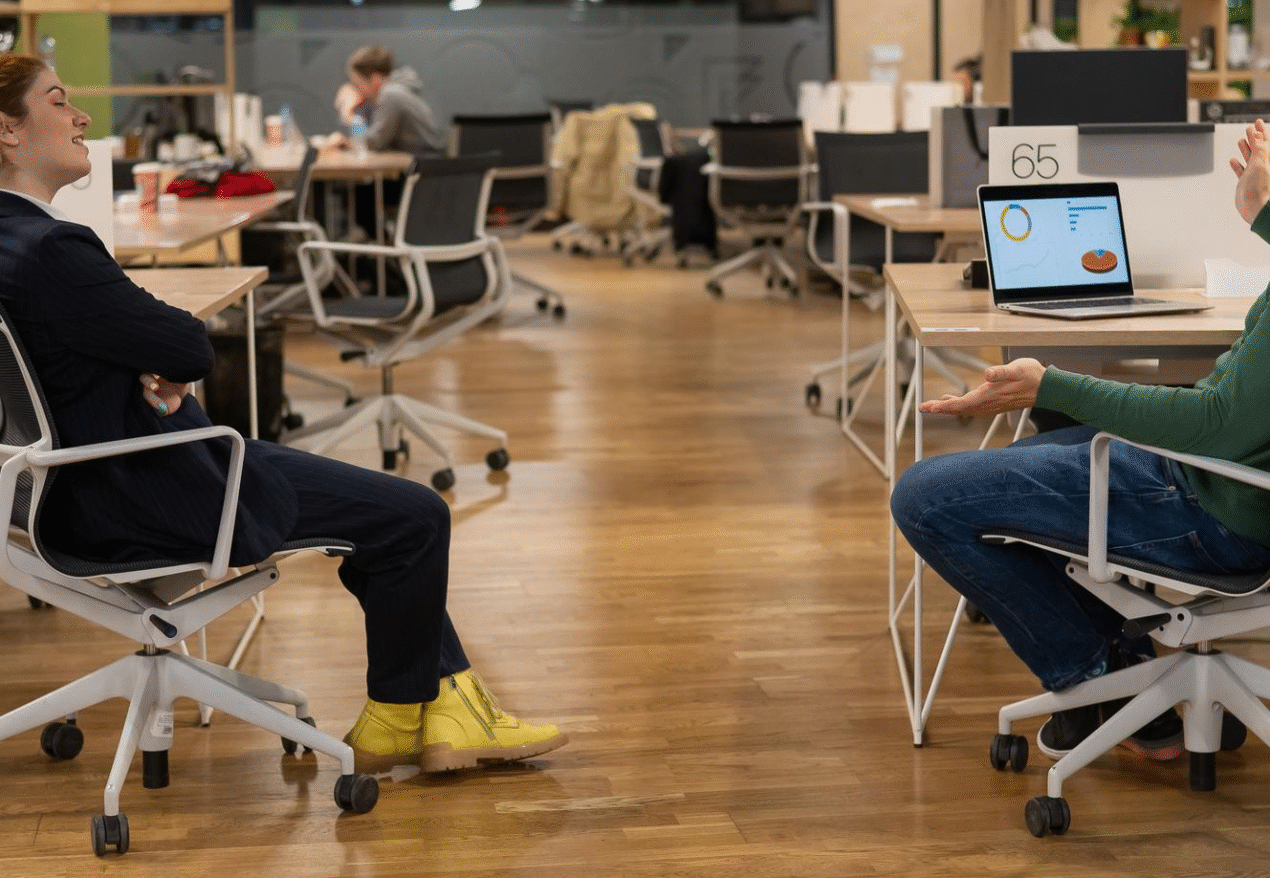 Two colleagues having a casual conversation in an open office space with modern workstations. One, a woman in a navy suit with yellow boots, is sitting back with a relaxed posture.