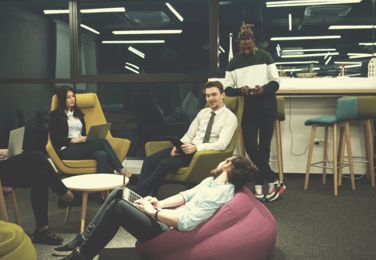 A modern office lounge with a casual setting where people are relaxing and conversing. Two individuals are sitting on bean bags playing video games, while three others are having a discussion on cozy armchairs. The background features a stylish kitchen area and vibrant decor.