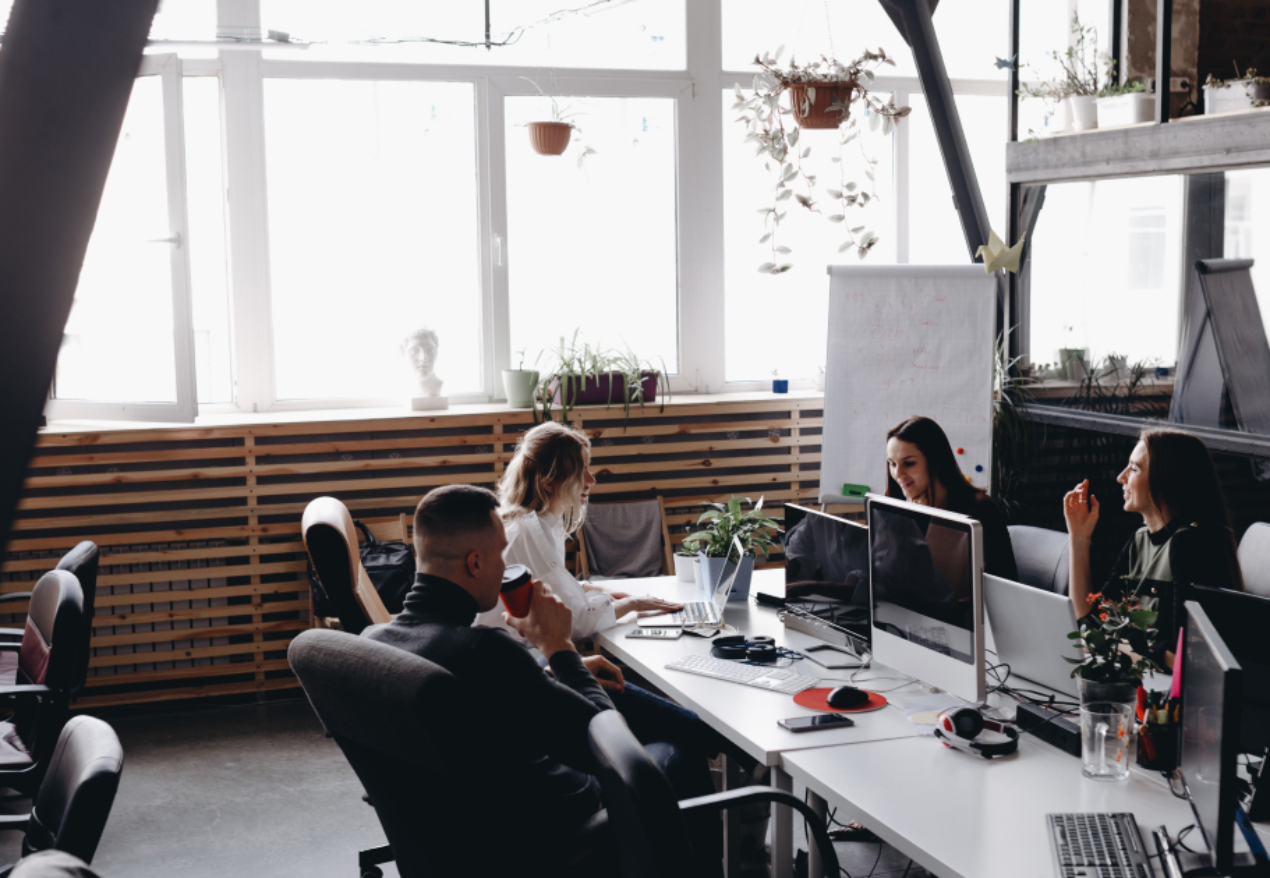 A modern office space with four colleagues working collaboratively at a shared desk. The room features large windows allowing natural light, indoor plants, and a mix of technology, including computers and monitors. A whiteboard is visible in the background.